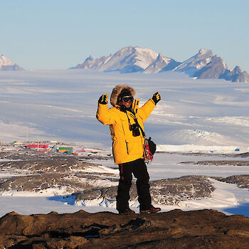 Expeditioner on top of a hillside overlooking sea ice, Mawson station and mountains in the distance.