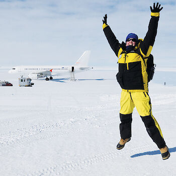 Yellow and black clad expeditioner jumping on the ice with A319 aircraft parked in background.