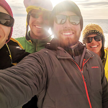 Team selfie of six people with sun and sea behind. All in assorted winter gear