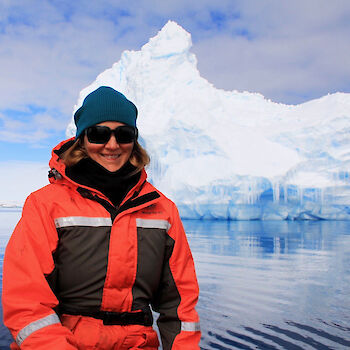 Expeditioner in red and black dry suit with blue beanie and black sun glasses and large iceberg in background.