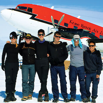 Group of six expeditioners standing with arms across each others shoulders, behind the front portion of a Basler aircaft painted white underneath and red on top.