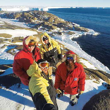 Group of four expeditioners in goose down jackets and goggles, standing on peninsula with sea and glacier in background.