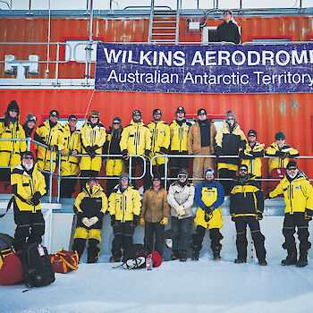 Group of expeditioners in Antarctic clothing standing in from of Wilkins Aerodrome building.