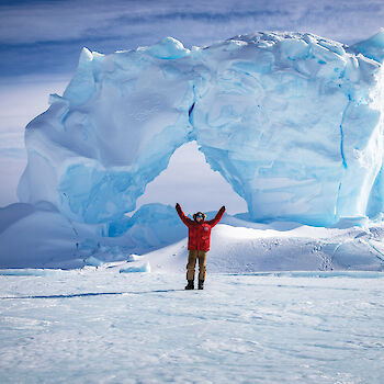 Smiling expeditioner standing in front of a glacier with arms raised up.
