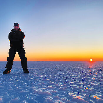 Station leader standing on the ice with the sunrise behind.
