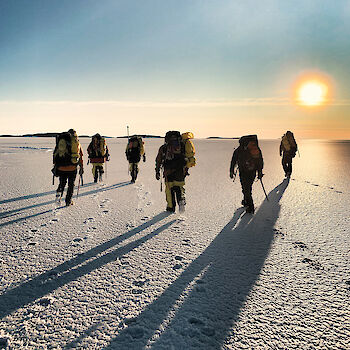 Group of expeditioners walking on the ice towards the sun at the horizon.