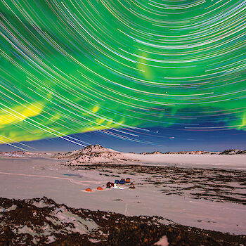 A small camp on the snow with a green aurora and star trails in the sky above.
