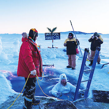 A man in a polar bear suit floats in an ice hole cut in to some sea ice whilst colleagues look on. Ice sculptures surround the ice hole and a home made sign saying 'Davis Pool ANARE 74 2021' has been erected beside the pool.