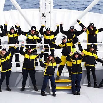A group of female scientists wave and smile to camera on the bow of the RV Investigator during the TEMPO voyage, all wearing the same cold weather jackets..