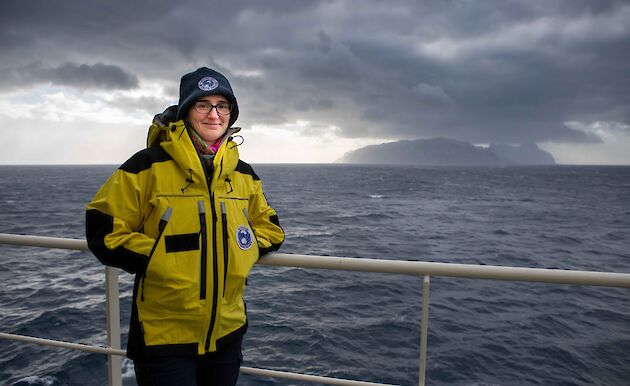 A woman stands in warm weather gear at the railings on the side of a ship. An island can be seen in the far distance.
