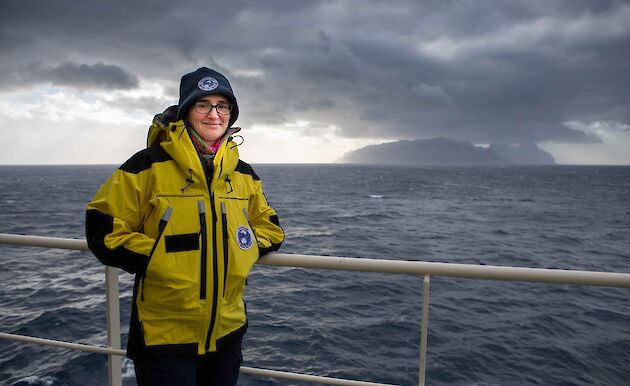 A woman stands in warm weather gear at the railings on the side of a ship.  An island can be seen in the far distance.