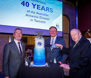 Two men standing next to a three tiered cake and one man holding a knife cutting the cake