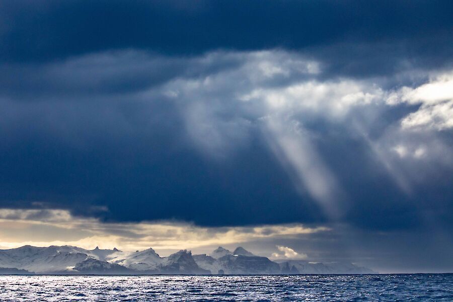 Sunlight streams through gathered dark clouds in the sky above a snow capped mountainous island.