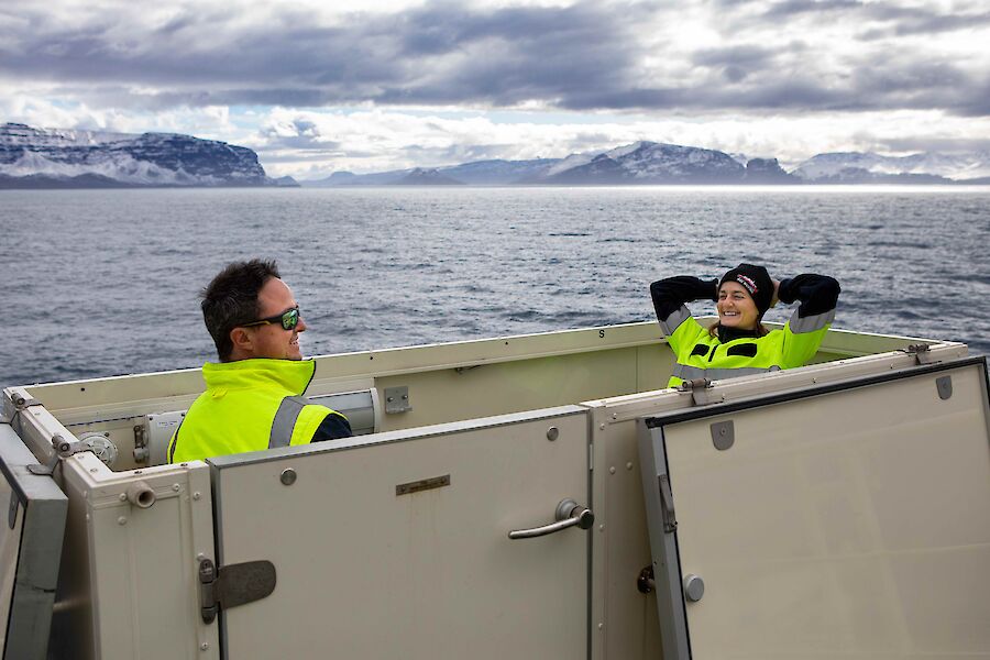 Two people in hi-vis sitting in a open top box at the back of a boat.  One person has their hands behind their head, leaning back and smiling.  Snowy mountains can be seen in the background.