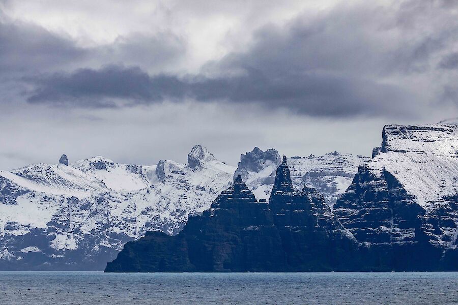 Looking from ship towards steep, dramatic cliff faces, with snowy mountains.