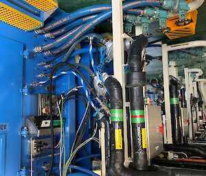 Blue shipping container lab connected to ship's services via umbilical pipes and cables.