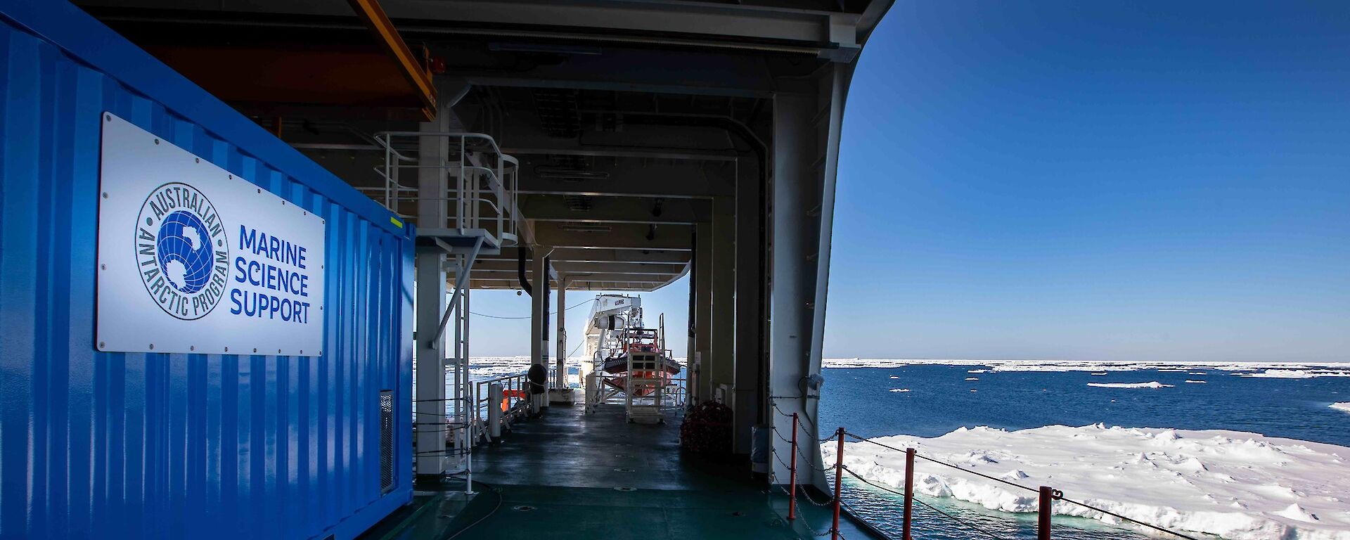 The deck of a ship as it sails through the ocean, surrounded by floating chunks of ice. The sky is clear and blue. A shipping container on the deck says 'Australian Antarctic Program: Marine Science Support'.