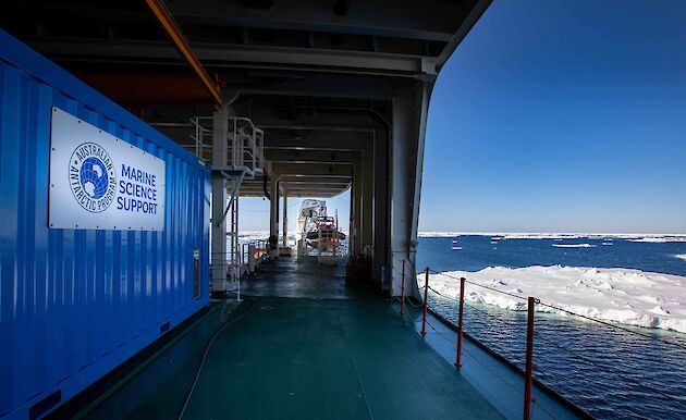 The deck of a ship as it sails through the ocean, surrounded by floating chunks of ice. The sky is clear and blue. A shipping container on the deck says 'Australian Antarctic Program: Marine Science Support'.