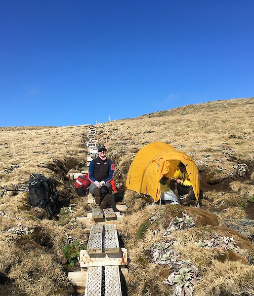 A yellow tent sits to the side of a rough wooden boardwalk on a hill with two people sitting down.