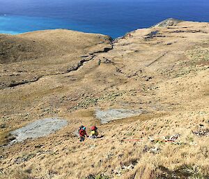 A view from the top of a hill looking out to sea with two people walking up the hill