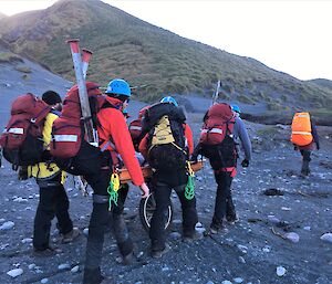 Five people with packs walk across a grey beach carrying a stretcher back to station