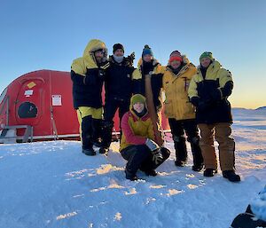 Six people in standing on the snow in front of a red hut.