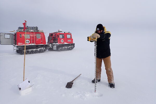A man drills the sea ice in front of a red vehicle