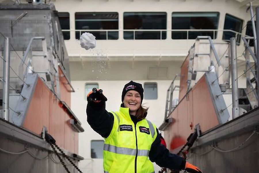 A woman in hi vis stands on the snowy dusted deck of a ship and throws a snowball at the camera