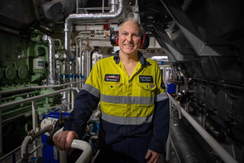 A man stands in hi-vis in the engine room of a ship smiling to camera