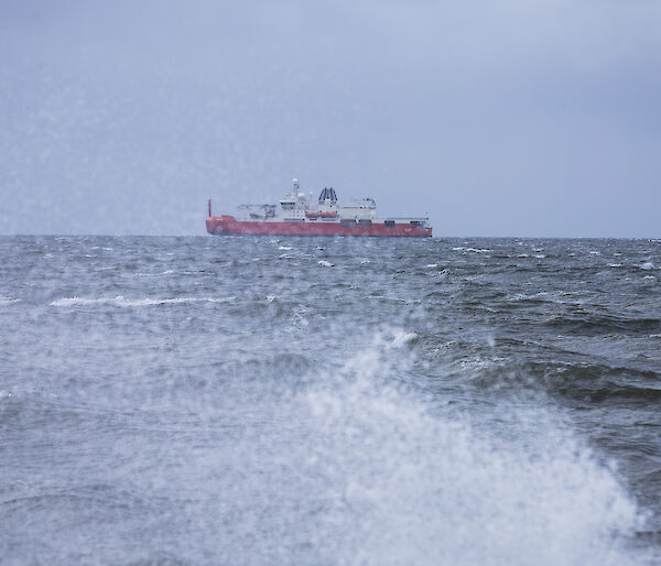 A red ship is seen from the shore behind a breaking wave