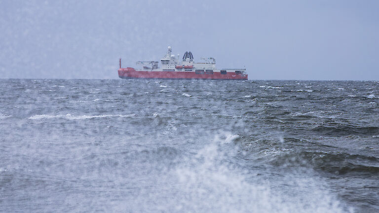 A red ship is seen from the shore behind a breaking wave