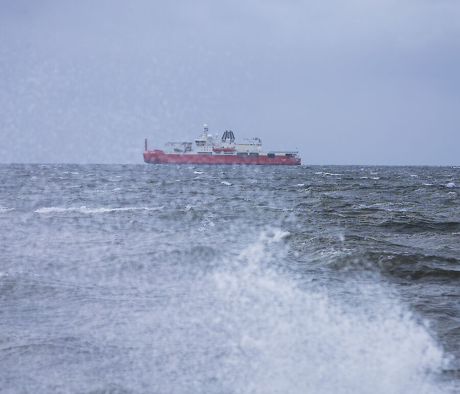 A red ship is seen from the shore behind a breaking wave