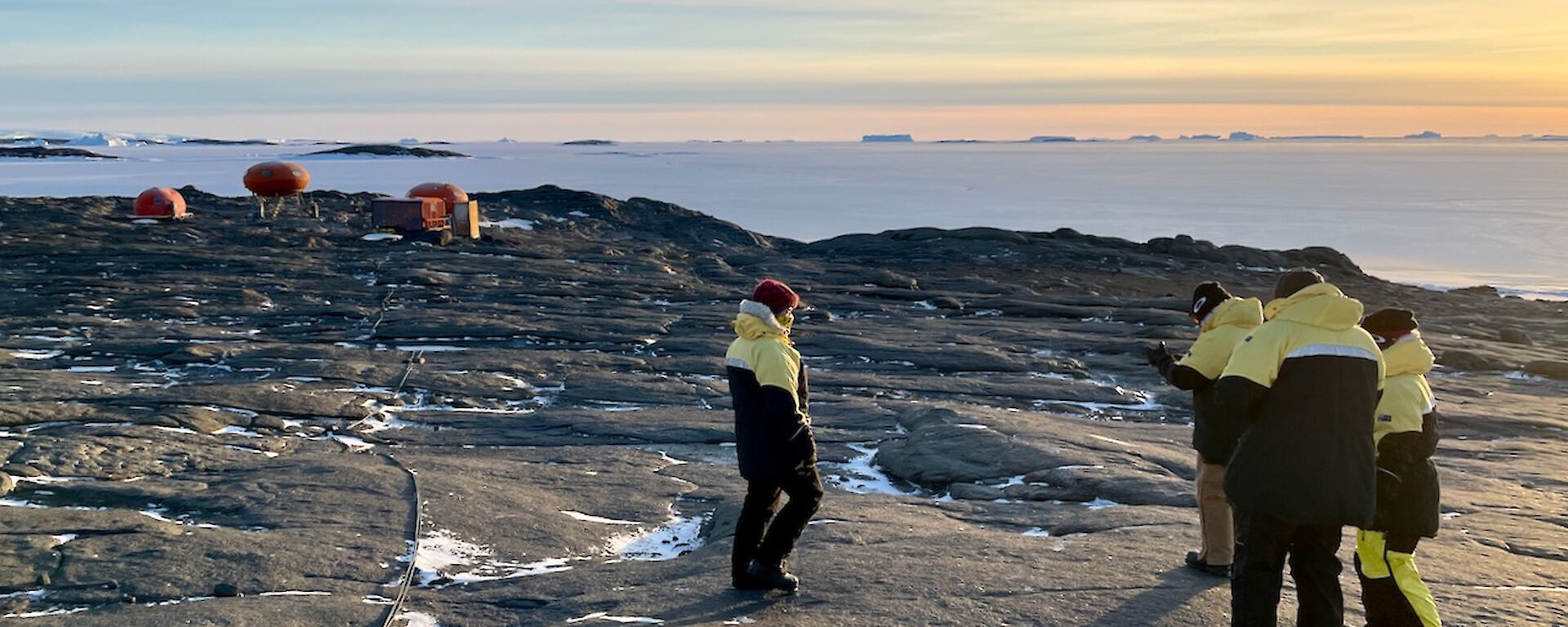 Four expeditioners in their black and yellow winter jackets with Béchervaise camp in the background.