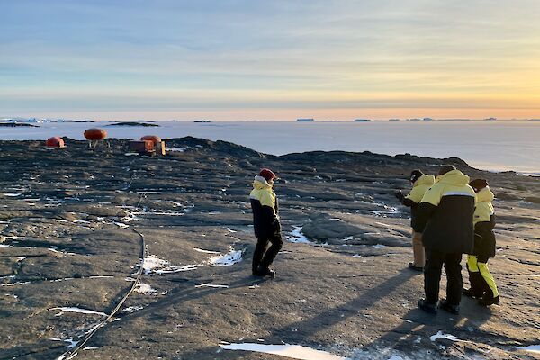 Four expeditioners in their black and yellow winter jackets with Béchervaise camp in the background.