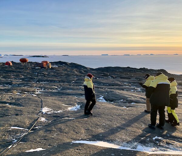 Four expeditioners in their black and yellow winter jackets with Béchervaise camp in the background.