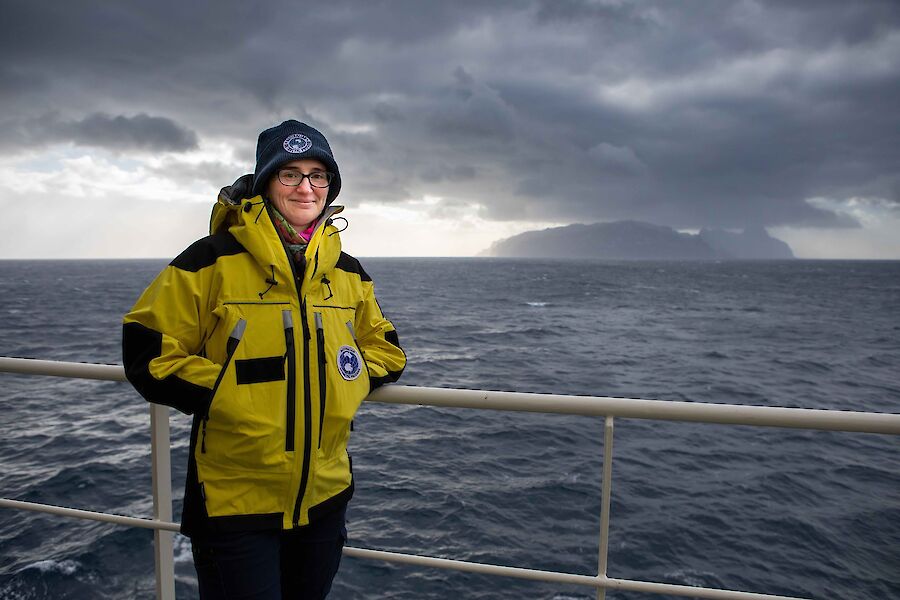 a woman stands at the rail of a ship in wet weather gear, smiling to camera, with an island visible on the horizon