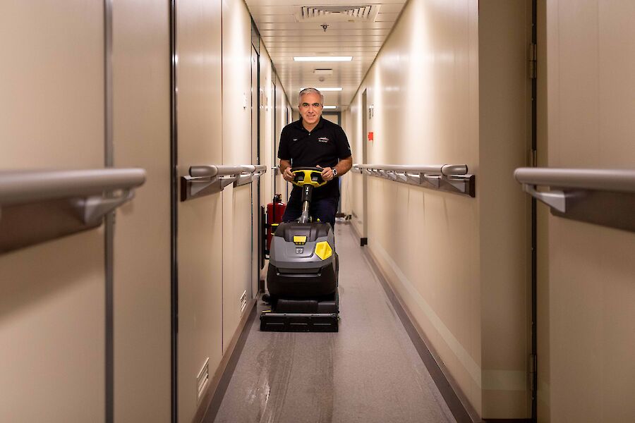 A man in a narrow ship corridor pushing a floor cleaning machine