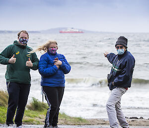 Three people giving the thumbs-up to the camera, with the RSV Nuyina in the background.