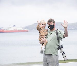 Man with camera holding a child and waving; RSV Nuyina in the background