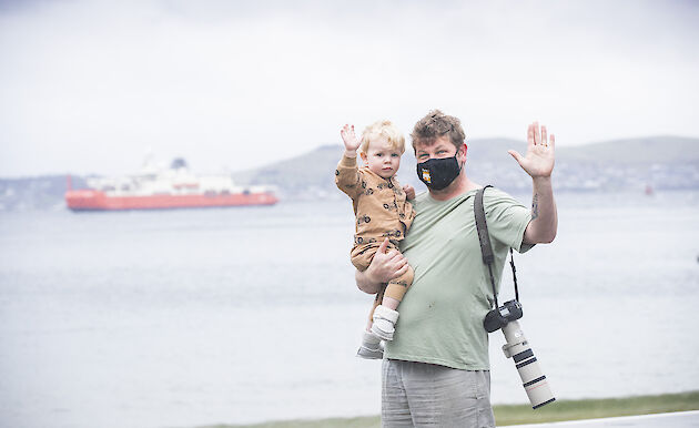 Man with camera holding a child and waving; RSV Nuyina in the background
