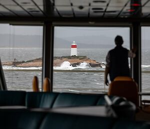 Silhouette of man standing in front of ship window looking out to a rock with a beacon on it and waves breaking around it.