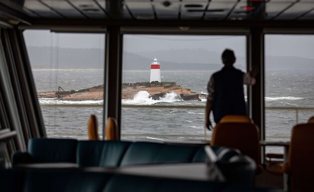 Silhouette of man standing in front of ship window looking out to a rock with a beacon on it and waves breaking around it.