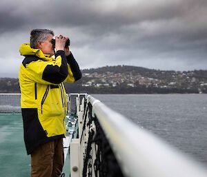 Man wearing yellow and black Antarctic issue jacket standing on deck of a ship peering through binoculars