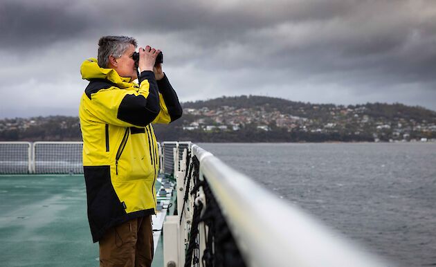 Man wearing yellow and black Antarctic issue jacket standing on deck of a ship peering through binoculars