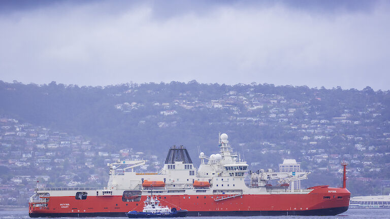 A large red and white ship sailing up river with houses visible on the hillside behind