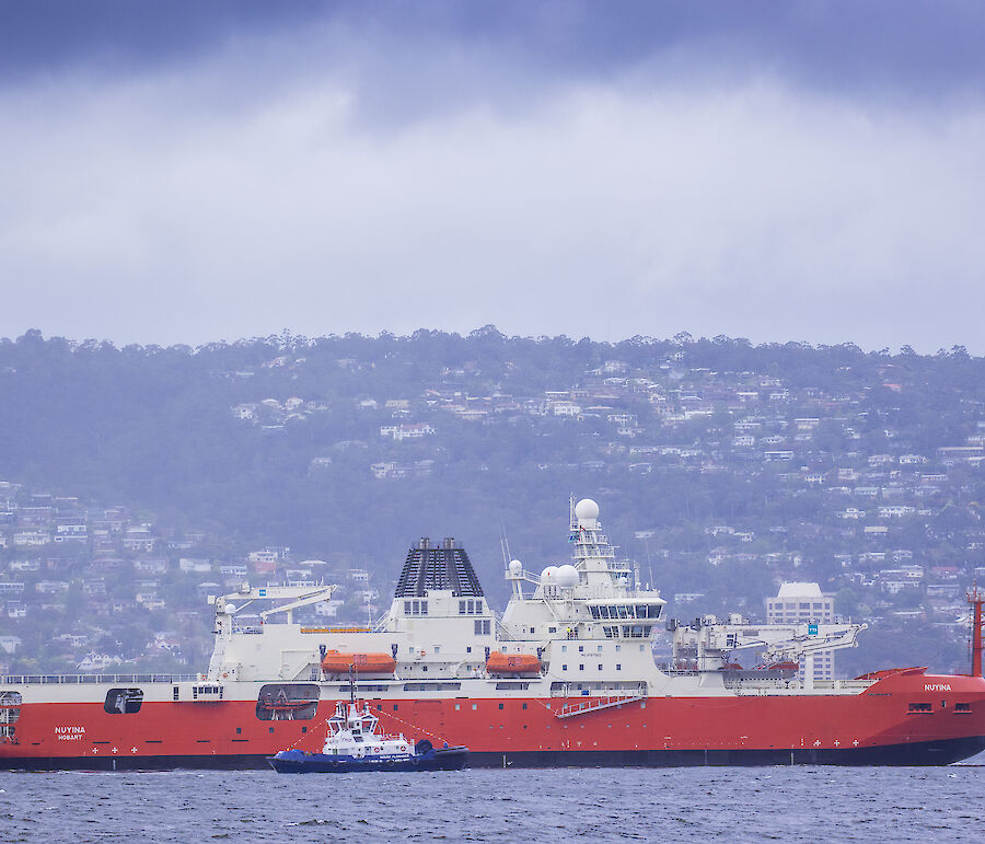 A large red and white ship sailing up river with houses visible on the hillside behind