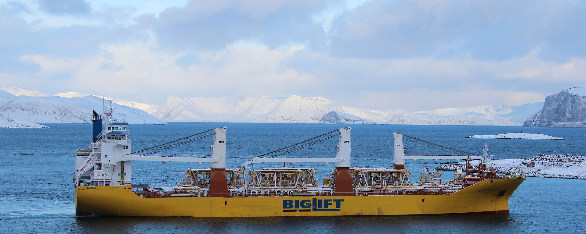 Large yellow and white cargo ship in a bay with snow capped mountains behind
