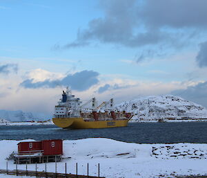 Large yellow and white cargo ship in a bay with snow capped mountains behind in front