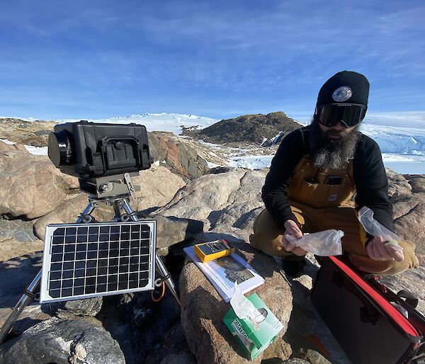 Expeditioner kneeling down working on rocks servicing remote penguin camera