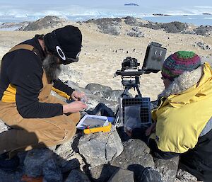 Two expeditioners kneeling down working on rocks servicing remote penguin camera